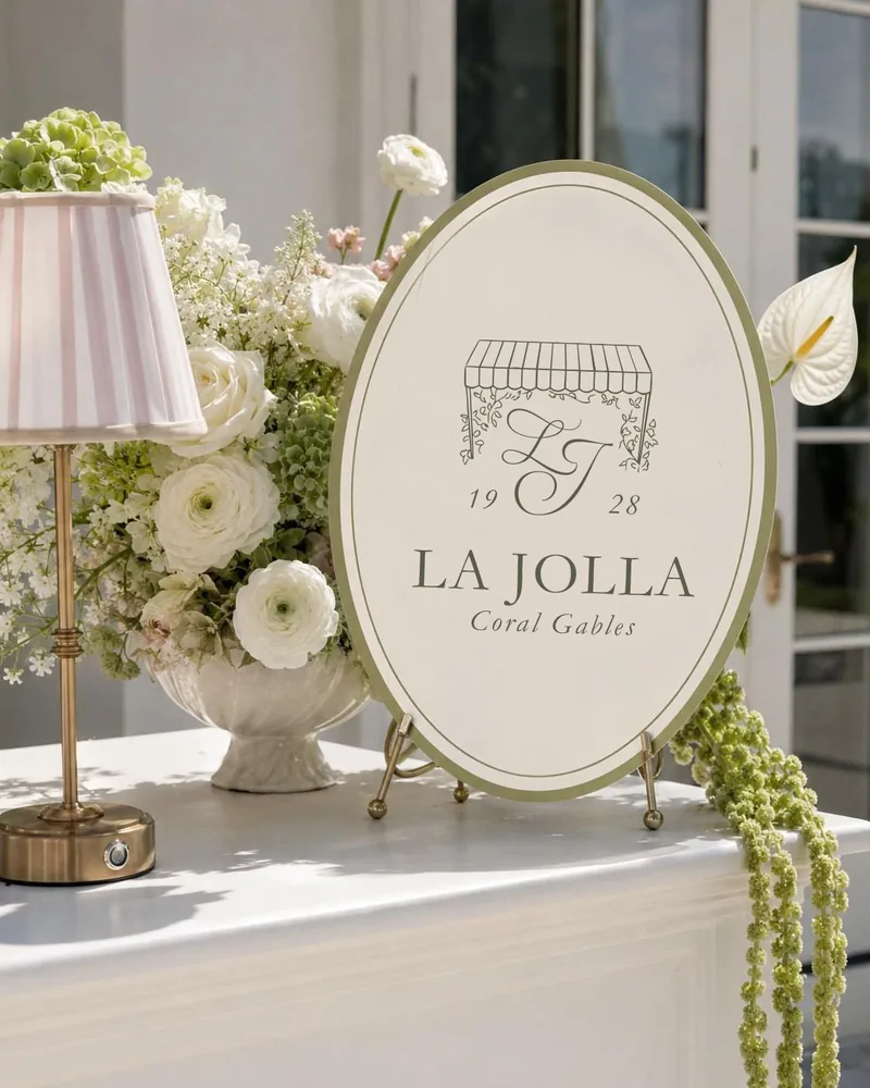 An oval La Jolla Coral Gables 1928 sign beside white flowers and a striped lamp.