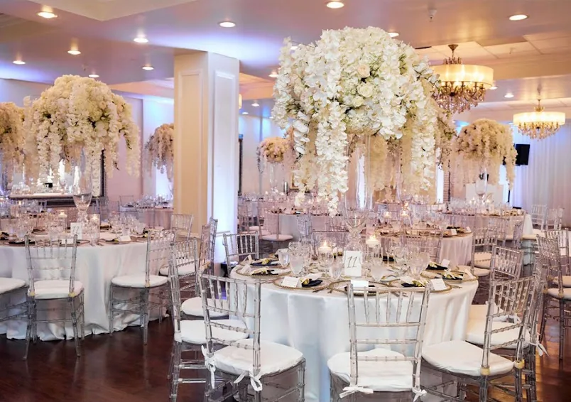 Round tables beneath towering white orchid and hydrangea centerpieces, with clear chairs and chandeliers.