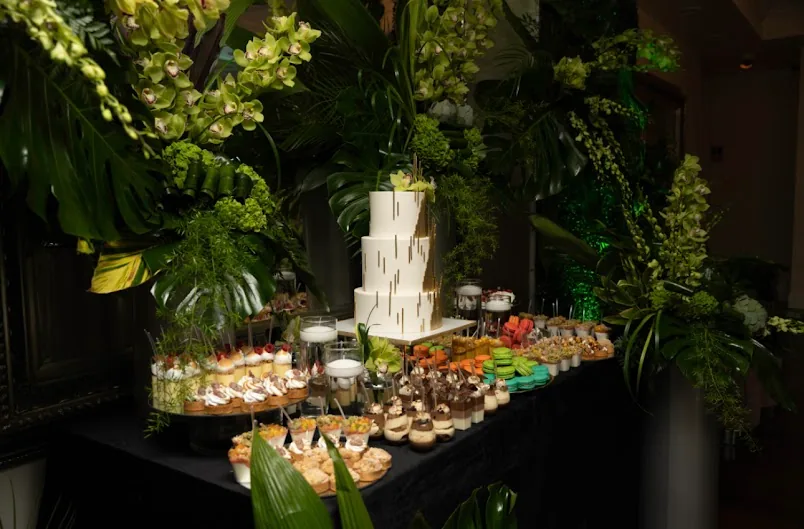 A dessert table with a tiered cake among tropical greenery.
